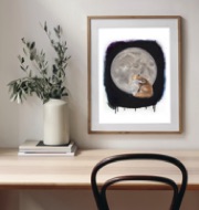 A realistic painting of a fox sitting in front of a full moon framed in a natural wood frame hung above a desk with a leafy plant in a vase sitting on books to the left and a chair in front.